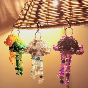 May include: Three beaded jellyfish ornaments in rainbow, clear, and purple hues hang from a lamp. The jellyfish have beaded bodies and dangling beaded tentacles. The rainbow jellyfish has orange, yellow, green, and blue beads. The clear jellyfish has white and blue beads. The purple jellyfish has pink and purple beads.