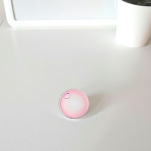 May include: A small, round, pink and white object with a clear border. A small, purple, circular design is on the top left of the object. The object is on a white surface.