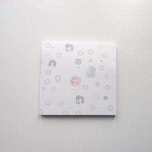Könnte beinhalten: Ein quadratischer Notizblock mit weißem Hintergrund, bedeckt mit einem Muster aus rosa, lila und pfirsichfarbenen Formen. Die Formen umfassen Kreise und abstrakte Formen, die ein wissenschaftliches oder biologisches Thema suggerieren.