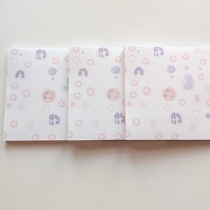 Könnte beinhalten: Drei quadratische weiße Karten mit einem sich wiederholenden Muster aus rosa und lila Formen. Die Formen umfassen Kreise, Bögen und eine stilisierte Figur. Die Karten sind nebeneinander auf einer weißen Oberfläche angeordnet.