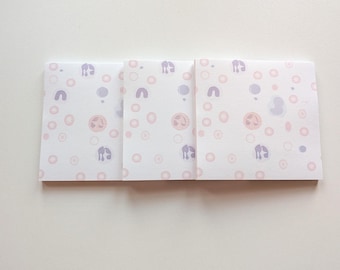 May include: Three white square cards with a repeating pattern of pink and purple shapes. The shapes include circles, arches, and a stylized figure. The cards are arranged side by side on a white surface.