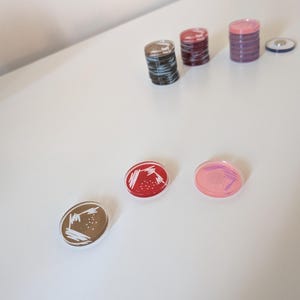 Könnte beinhalten: Eine Sammlung farbenfroher, runder Magnete mit verschiedenen Designs. Die Magnete umfassen Stapel zylindrischer Magnete in Schwarz-, Rot- und Lilatönen. Einzelne Magnete weisen braune, rote und rosa Hintergründe mit weißen Linienmustern auf.
