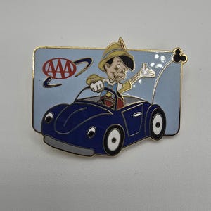 May include: Enamel pin featuring Pinocchio driving a blue car. The pin has a light blue background with the AAA logo. Pinocchio is wearing a yellow hat and waving. The car has white wheels and a gold border.