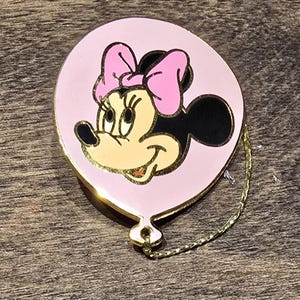 May include: A pink Minnie Mouse pin in the shape of a balloon. The pin features Minnie's face with a pink bow, black ears, and a gold outline. A gold chain hangs from the bottom of the balloon.