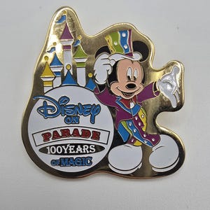 May include: A gold-colored Disney pin featuring Mickey Mouse in a purple and blue outfit, with a top hat. The pin has a castle and the words "Disney on Parade 100 Years of Magic."