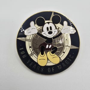 May include: A round pin featuring Mickey Mouse with his arms raised. The pin has a dark blue border with the text "100 YEARS OF MAGIC." Mickey is in his classic outfit, standing in front of a globe.