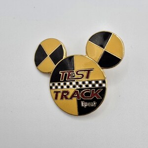 May include: A gold-colored pin in the shape of Mickey Mouse ears, with the words "TEST TRACK" and "Epcot" on the front. The pin has a black and yellow color scheme, with a checkered pattern.