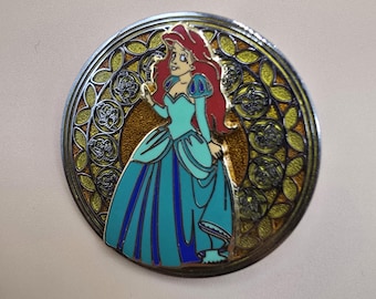 Pin oficial vintage de Disney que muestra a Ariel de La Sirenita con un vestido verde y azul sobre un fondo de vidrieras amarillas.