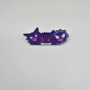 May include: A purple and black pin featuring three cartoon characters with various expressions. The central character has a wide grin with red eyes, flanked by two other characters with different facial expressions. The pin is on a white background.