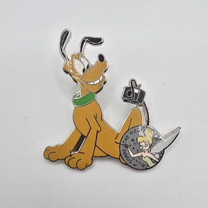 May include: A Pluto and Tinkerbell enamel pin. Pluto is depicted in a seated position, with a green collar. Tinkerbell is on a silver coin with the words "SHARE THE MAGIC". The pin has a black camera and silver accents.