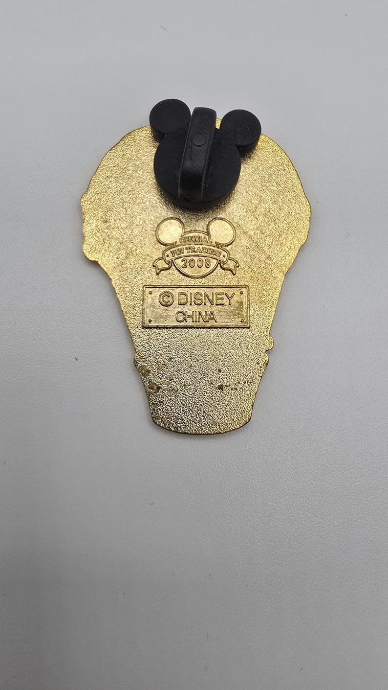 May include: A gold-toned metal pin back featuring a black Mickey Mouse-shaped rubber clutch. The back is embossed with the text Official Pin Trading 2008 and a rectangular box containing the text Disney China, indicating its origin and production year.