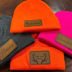Peut inclure: Quatre bonnets de couleurs variées : gris, orange et rose vif. Chaque bonnet est orné d'un écusson rectangulaire en cuir avec un nom ou une phrase. Le bonnet orange porte l'inscription "Dads Little Buck". Les autres bonnets portent les noms "Blaine", "Karter" et "Kendal".