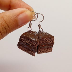 May include: A pair of novelty earrings resembling slices of chocolate cake. The earrings have a layered design with a dark chocolate topping and silver-coloured hooks.