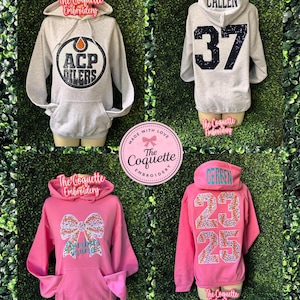 May include: Four hoodies on display. Two are gray, one with "ACP OILERS" and the other with "CALLEN 37". The other two are pink, one with a bow design and the other with "GERBEN 23 25". All have a front pocket.