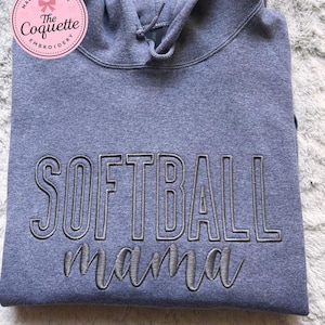 May include: A gray hooded sweatshirt with the embroidered text "SOFTBALL mama" in a contrasting gray color. The sweatshirt has a drawstring hood and a soft, textured appearance. A pink circular logo with text is in the upper left corner.