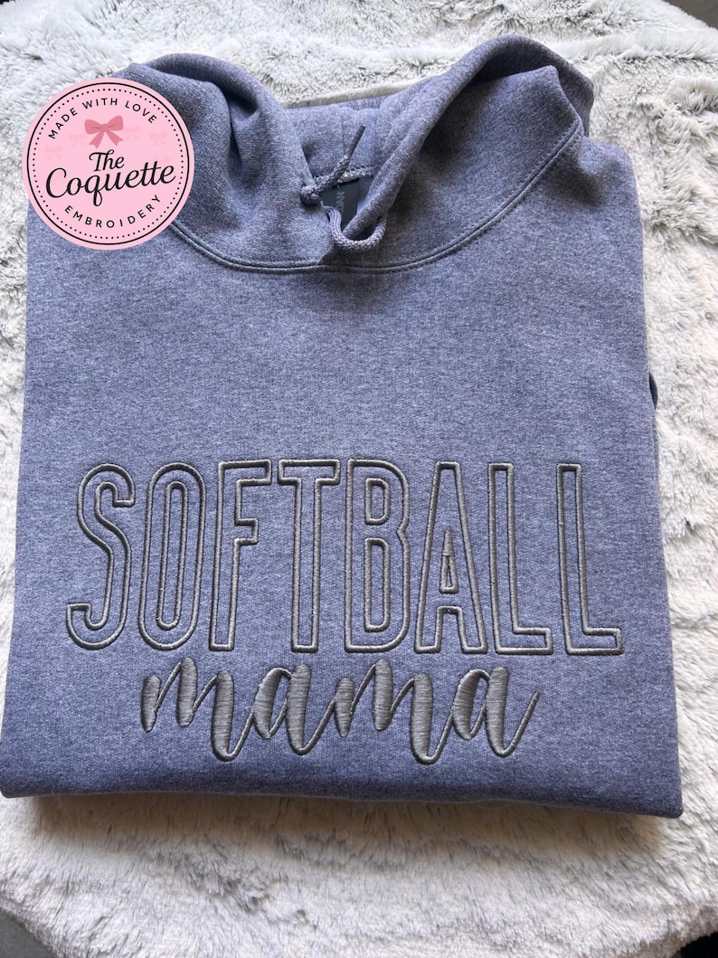 May include: A gray hooded sweatshirt with the embroidered text "SOFTBALL mama" in a contrasting gray color. The sweatshirt has a drawstring hood and a soft, textured appearance. A pink circular logo with text is in the upper left corner.
