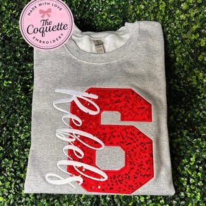 May include: A gray sweatshirt with a large red sequined number 6 and the word "Rebels" in white script. The sweatshirt has a crew neck and is folded. A pink and white circular sticker with text is in the upper left corner.