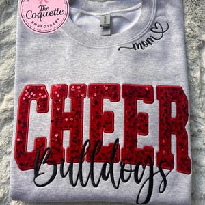 May include: A gray sweatshirt with the word "CHEER" in large red sequined letters and the word "Bulldogs" in black script. The word "mom" is embroidered in black near the neckline. A pink and white circular logo is in the upper left corner.