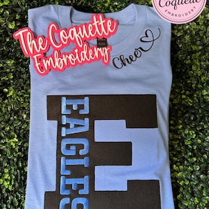 May include: A light blue t-shirt with the word "EAGLES" vertically embroidered in blue and black. The word "Cheer" is written in black script. The Coquette Embroidery logo is visible.