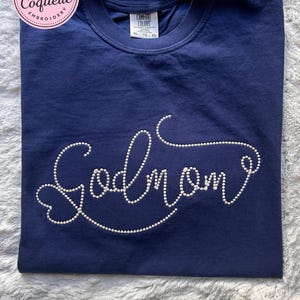 May include: Navy blue t-shirt with the word "Godmom" spelled out in white, dotted lettering. The shirt is a Comfort Colors brand, size XL-TG-EG. A pink and white circular logo with text is in the upper left corner.