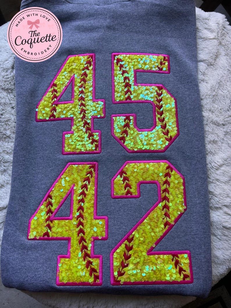 May include: Gray sweatshirt with the numbers 45 and 42 in a softball design. The numbers are yellow with red stitching and outlined in pink. The sweatshirt has a circular logo in the upper left corner that says "Made with Love" and "The Coquette Embroidery".