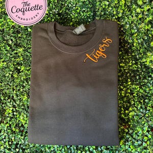 May include: A black crewneck sweatshirt featuring the word "tigers" embroidered in orange cursive script on the upper right chest. A circular pink logo with the text "MADE WITH LOVE The Coquette Embroidery" is positioned in the top left corner.