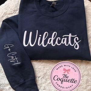 May include: Navy blue sweatshirt with the word "Wildcats" embroidered in white script across the chest. The left sleeve has "Go Cats!" embroidered in white. A pink and white circular logo is in the lower right corner.