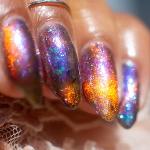 May include: Long, almond-shaped fingernails coated in a multi-dimensional nail polish featuring a deep purple base with shifting orange, gold, and teal iridescent shimmer, accented by fine holographic glitter particles throughout the finish.