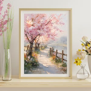 May include: A framed watercolor painting depicts a path along a river lined with blossoming pink cherry trees. A wooden fence borders the path. The painting is in a gold frame and is displayed on a wooden shelf with vases of flowers.