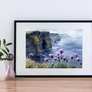 May include: A framed watercolor print of the Cliffs of Moher in Ireland. The artwork depicts the rugged cliffs, the ocean, and purple wildflowers in the foreground. The print is in a black frame and rests on a wooden surface.