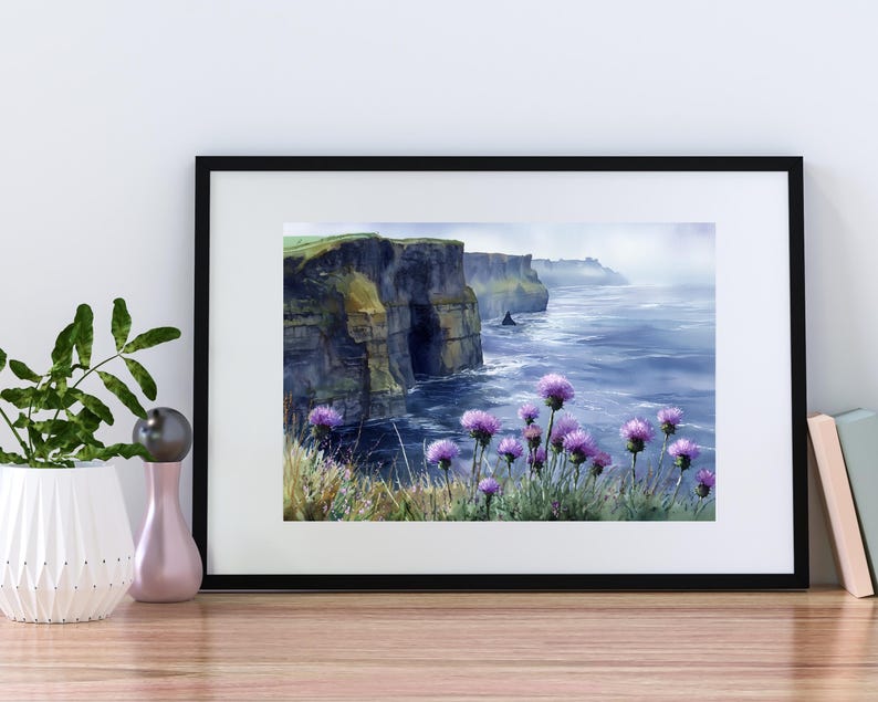 May include: A framed watercolor print of the Cliffs of Moher in Ireland. The artwork depicts the rugged cliffs, the ocean, and purple wildflowers in the foreground. The print is in a black frame and rests on a wooden surface.