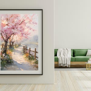 May include: A framed watercolor print depicts a pathway along a body of water, lined with blossoming pink cherry trees. A wooden fence borders the path. The artwork is displayed on a white wall, with a green sofa and wooden floor in the background.