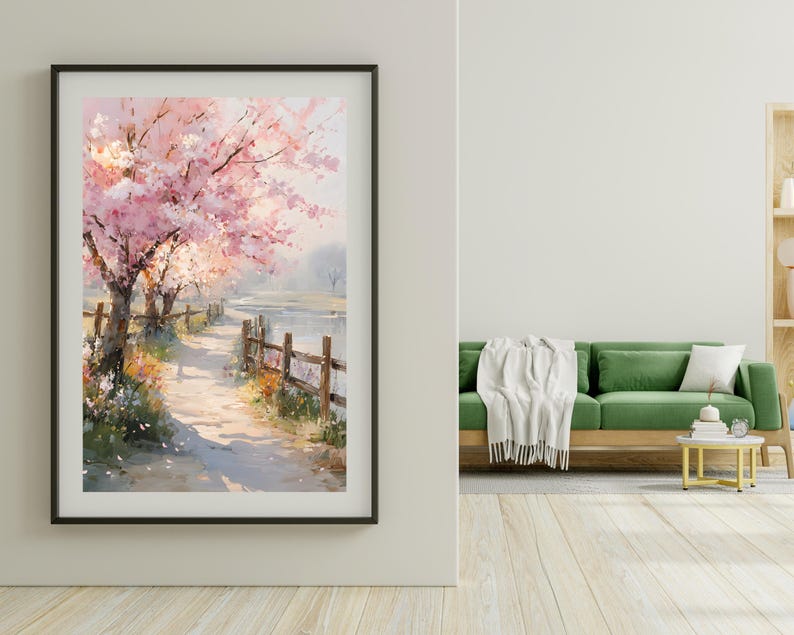 May include: A framed watercolor print depicts a pathway along a body of water, lined with blossoming pink cherry trees. A wooden fence borders the path. The artwork is displayed on a white wall, with a green sofa and wooden floor in the background.