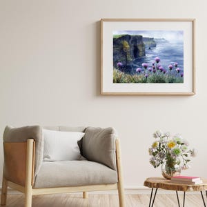 May include: A framed watercolor print of a coastal landscape featuring dramatic cliffs, the ocean, and purple wildflowers. The artwork is displayed in a light-colored wooden frame, hanging on a neutral-toned wall. The scene evokes a sense of tranquility.