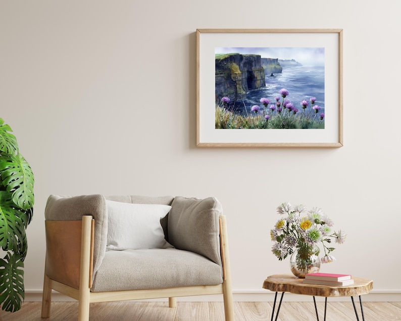 May include: A framed watercolor print of a coastal landscape featuring dramatic cliffs, the ocean, and purple wildflowers. The artwork is displayed in a light-colored wooden frame, hanging on a neutral-toned wall. The scene evokes a sense of tranquility.