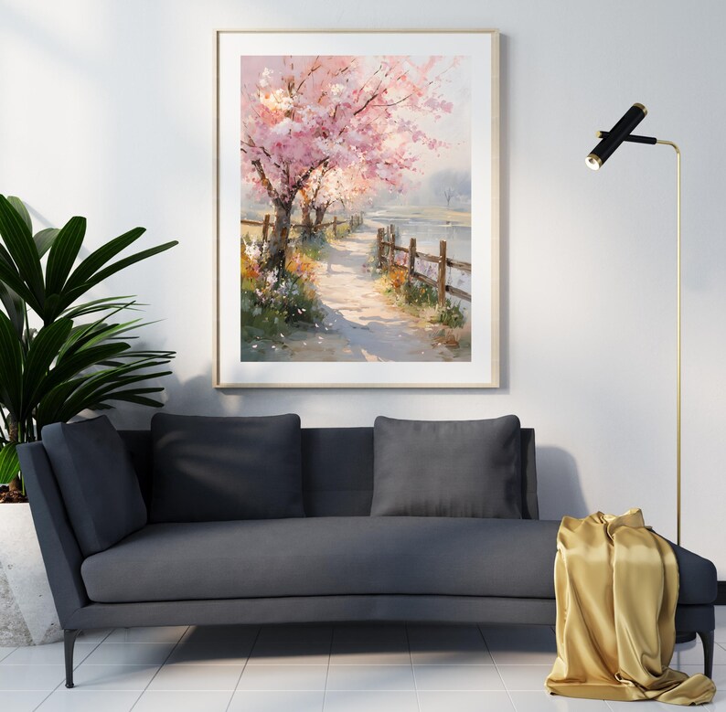 May include: A framed watercolor painting depicts a serene pathway lined with blossoming pink cherry trees. A wooden fence borders the path, leading to a body of water. The artwork is displayed above a dark gray sofa with pillows.