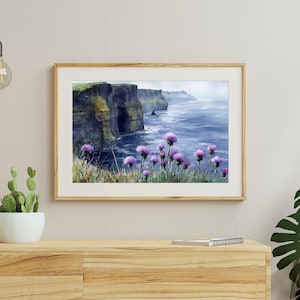 May include: A framed watercolor painting depicts a coastal landscape with dramatic cliffs and the ocean. Purple wildflowers bloom in the foreground. The artwork is displayed on a wooden cabinet, with a cactus and decorative lights.