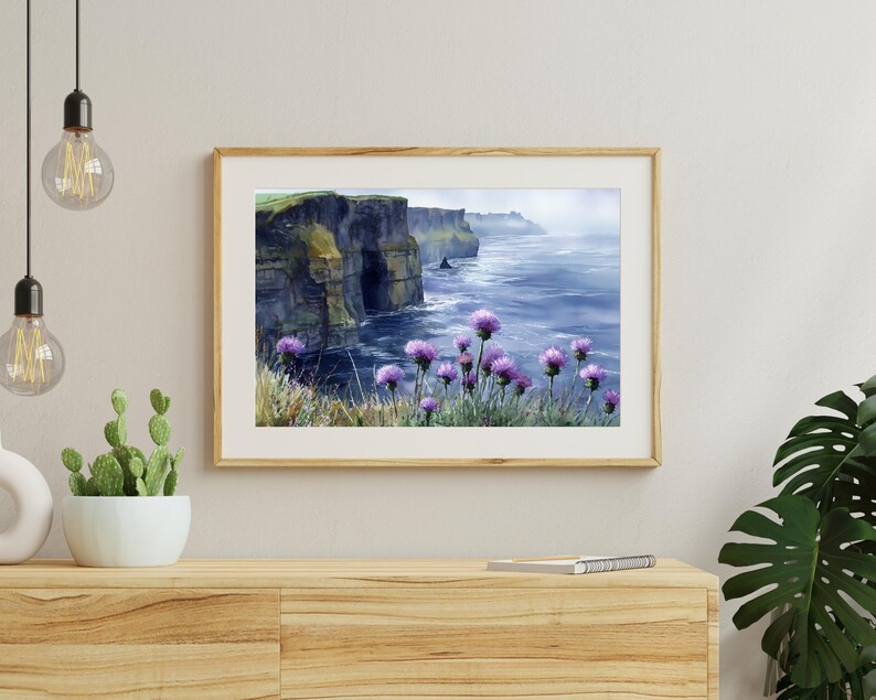 May include: A framed watercolor painting depicts a coastal landscape with dramatic cliffs and the ocean. Purple wildflowers bloom in the foreground. The artwork is displayed on a wooden cabinet, with a cactus and decorative lights.