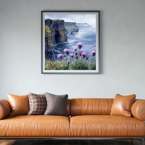 May include: Framed watercolor print of a coastal landscape featuring dramatic cliffs and the ocean. Purple wildflowers bloom in the foreground. The artwork is displayed above a brown leather sofa with decorative pillows.