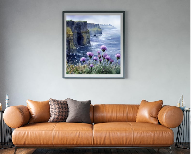 May include: Framed watercolor print of a coastal landscape featuring dramatic cliffs and the ocean. Purple wildflowers bloom in the foreground. The artwork is displayed above a brown leather sofa with decorative pillows.