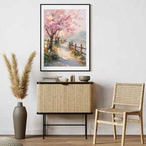 May include: A framed watercolor painting depicts a serene landscape with a path alongside a body of water. Pink cherry blossom trees line the path, and a wooden fence borders the water. The painting is displayed above a wooden cabinet and a woven chair.