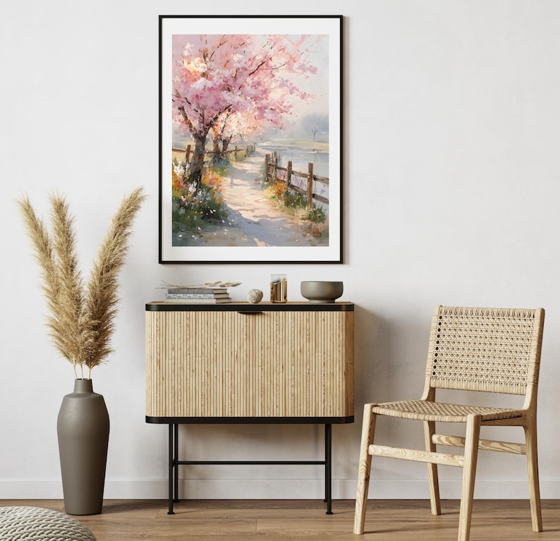 May include: A framed watercolor painting depicts a serene landscape with a path alongside a body of water. Pink cherry blossom trees line the path, and a wooden fence borders the water. The painting is displayed above a wooden cabinet and a woven chair.
