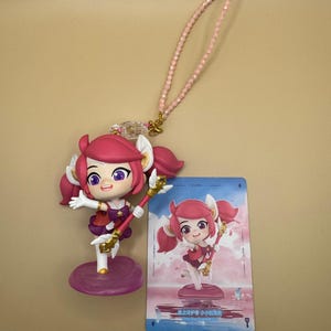 May include: A character figurine with pink hair and a staff, on a purple base. A card with a similar illustration is next to it. A pink beaded strap is attached to the figurine.
