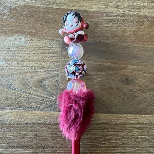 May include: A decorative pen with a burgundy barrel and silver tip. The pen is adorned with a sleeping doll figurine, a clear iridescent bead, a cluster of blue and pink roses, and a fluffy burgundy feather accent.