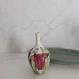 May include: A small, hand-painted ceramic vase with a bird design. The vase features a white base with a maroon bird, orange beak, and green and black accents. The vase is approximately 6 inches tall.