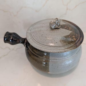 May include: A ceramic pot with a side handle and a matching lid featuring a small sculpted knob. The pot has a two-tone glaze, with one side in a dark brown and the other in a speckled light gray. The lid is also speckled light gray with brown accents.