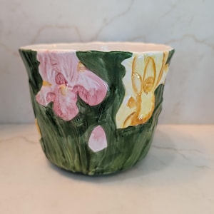 May include: A ceramic planter featuring a raised, three-dimensional floral design. The exterior is painted with dark green foliage and large, prominent iris flowers in shades of soft pink and pale yellow, with a smooth, glossy finish throughout.