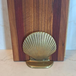 May include: A wooden cutting board with a brass seashell-shaped holder. The cutting board is made of various wood tones, including reddish-brown and light brown. The brass holder is in the foreground, adding a decorative touch to the kitchen item.