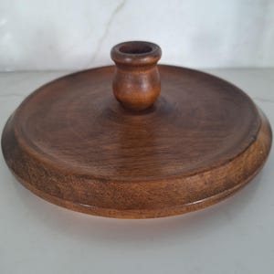 May include: A round, dark brown wooden candle holder with a small, cylindrical cup in the center. The holder has a wide, flat base and a smooth, polished finish. The wood grain is visible.