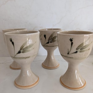 May include: Four handmade ceramic goblets with a beige glaze and a floral design in black and green. The goblets have a rounded base and a stem, perfect for serving beverages.